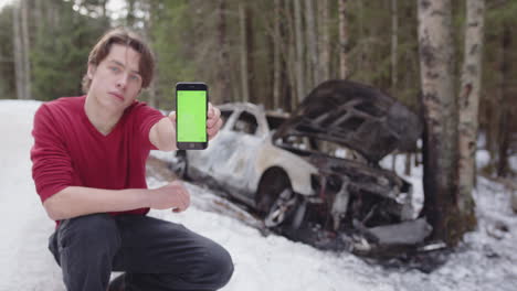 guy near wrecked car next to icy winter road holds out phone with green screen