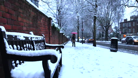 snowy london city scene with snow piled up on a bench and person with a red umbrella