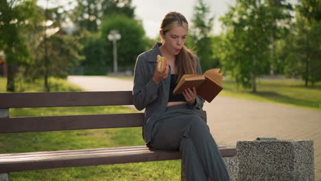 dama comiendo un bocadillo mientras lee un libro al aire libre, sentada en un banco del parque con las piernas cruzadas, disfrutando de un tiempo tranquilo en el parque verde, rodeada de árboles, un camino y un edificio en el fondo