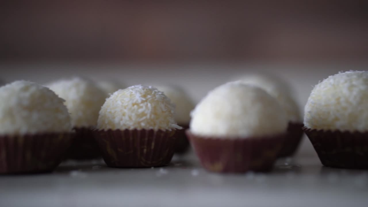 bolas blancas dulces cubiertas de coco seco colocadas en pequeños forros de papel marrón en la encimera, bandeja a la derecha