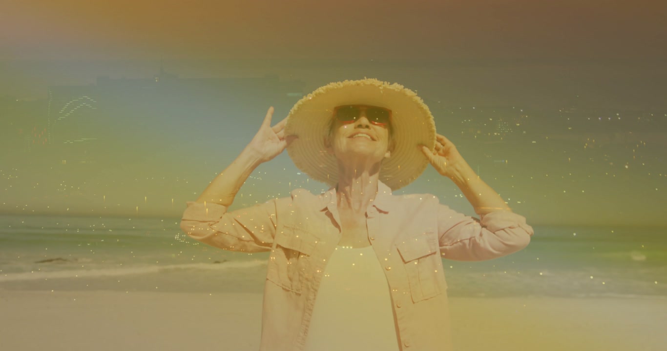 Senior woman feeling warm sun, holding hat, adjusting brim on beach, causing sparkles for travel