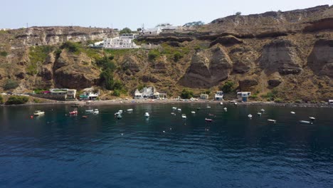 Aerial-flying-towards-Greek-Island,-rocky-cliffs-with-villas-and-fishing-boats-in-the-Mediterranean-Sea-in-Santorini,-Greece