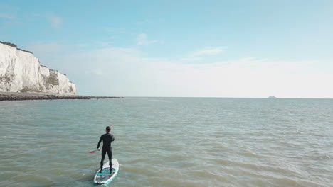 un joven se pone de pie haciendo paddle surf en el mar con los acantilados blancos de dover y el cielo azul de fondo