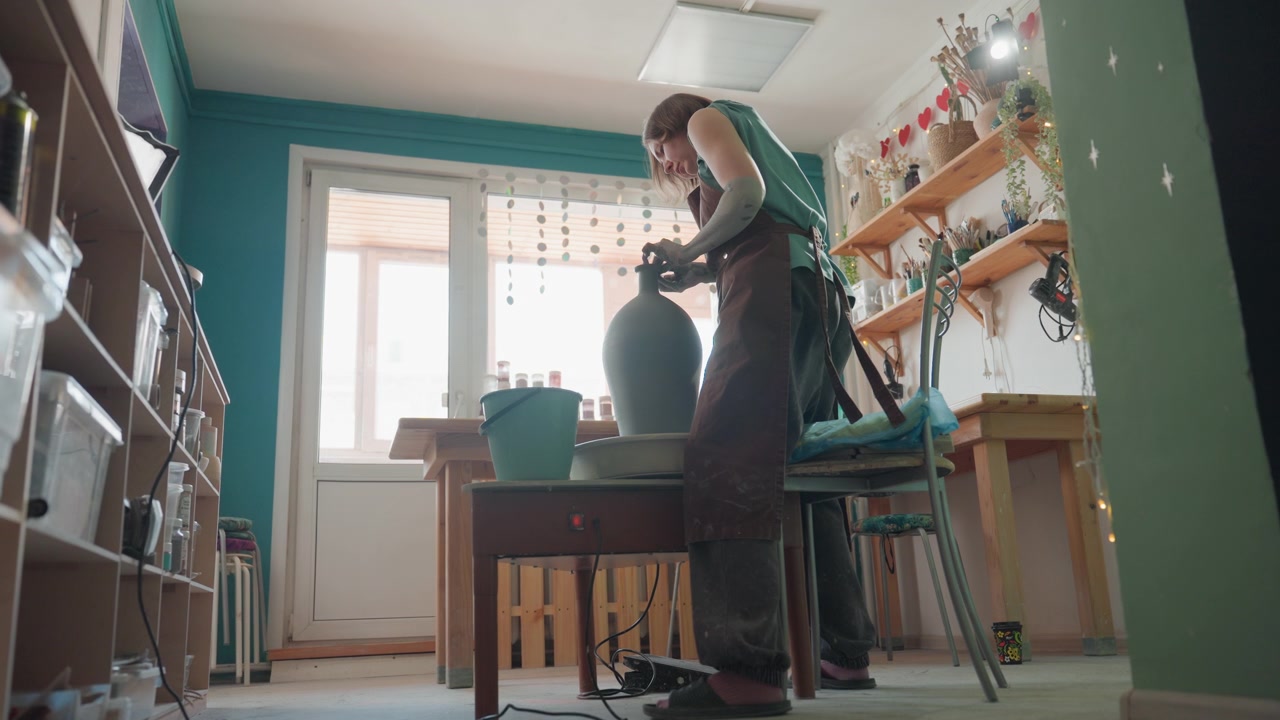 En vit kvinna färdigställer en hög vas på drejskivan i en ljus keramikverkstad, stående vid arbetsbänken med händerna som slätar ut formen. Hyllor med keramik och solljus strömmar in genom fönstret, vilket skapar en lugn och stolt stämning med en taktil slutkänsla.
