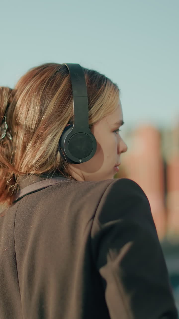 Lady in black suit enjoys music with eyes closed under warm sunlight ambiance, standing outdoors with building and parked cars in background