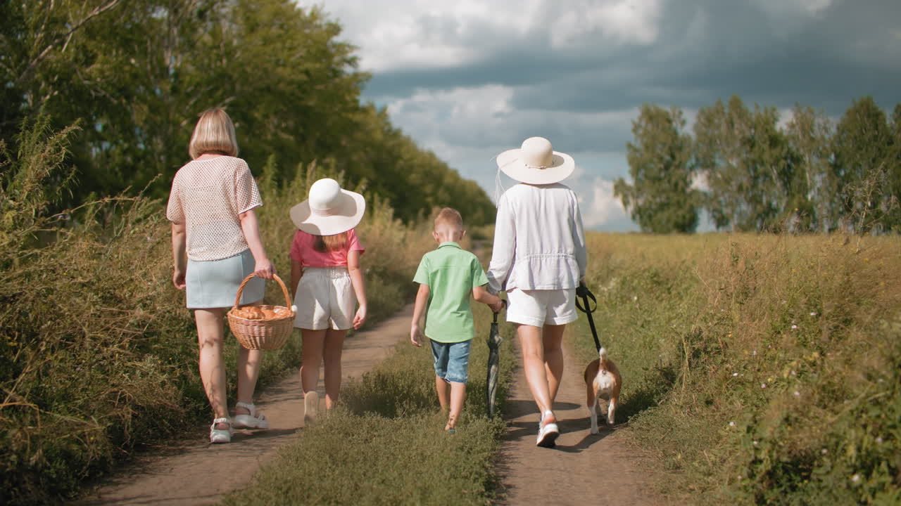 울창한 초록색으로 둘러싸인 길을 고 있는 가족의 뒷면, 피크닉을 하러 가는 동안 피크닉 바구니를 들고 있으며, 아이는 부분적으로 구름이 가득한 하늘 아래 우산을 들고 개를 줄에 고 있습니다.