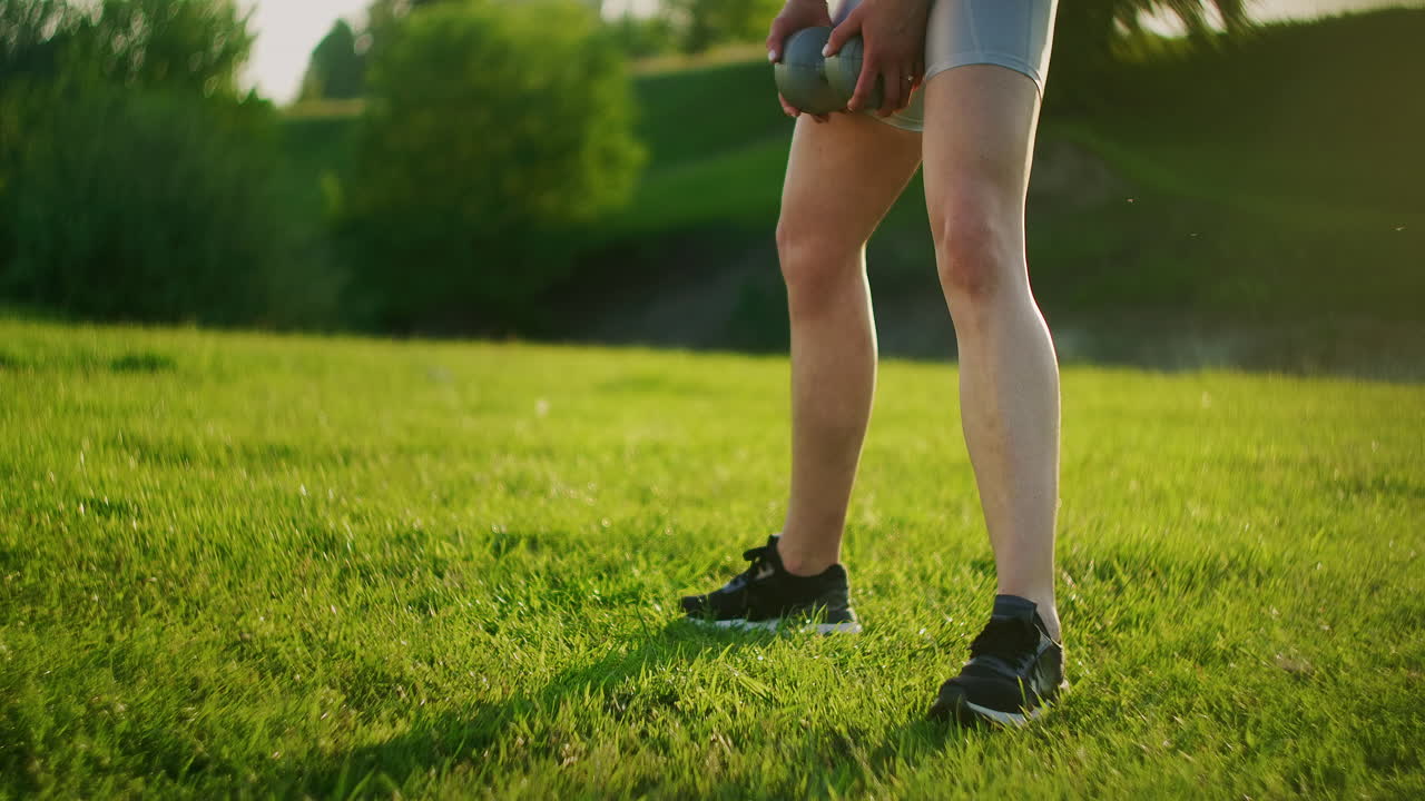 Close-up of a squat woman's leg in a park on grass with dumbbells