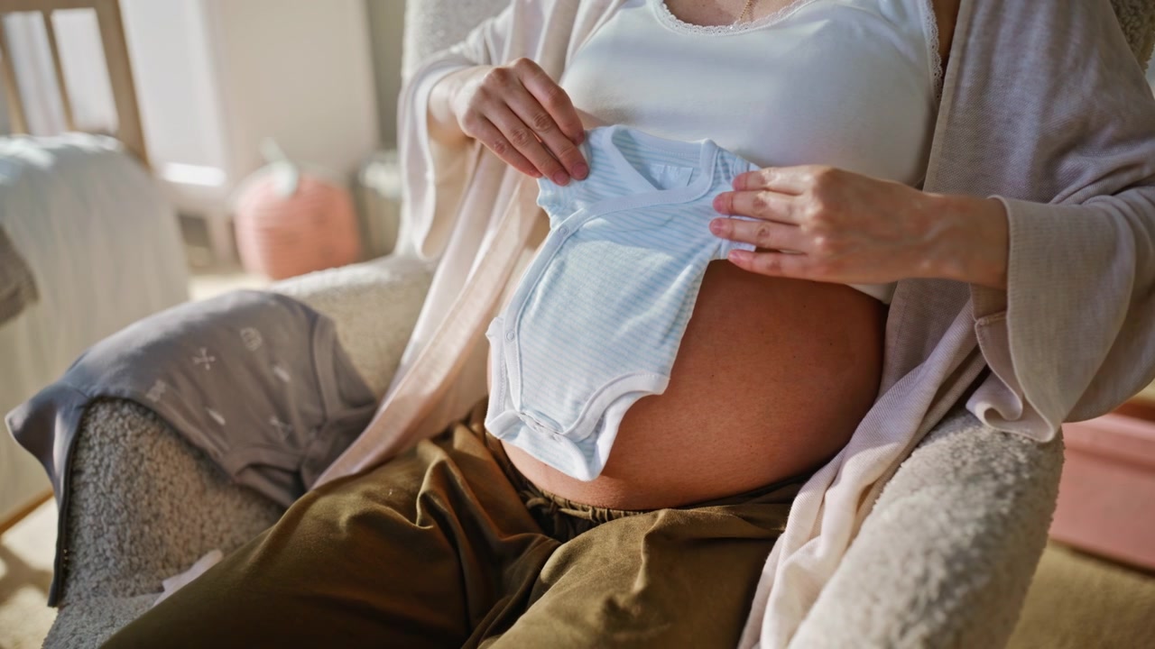 Donna incinta che tiene un body blu per neonato sulla pancia. Mamma sorridente, cameretta per neonati.