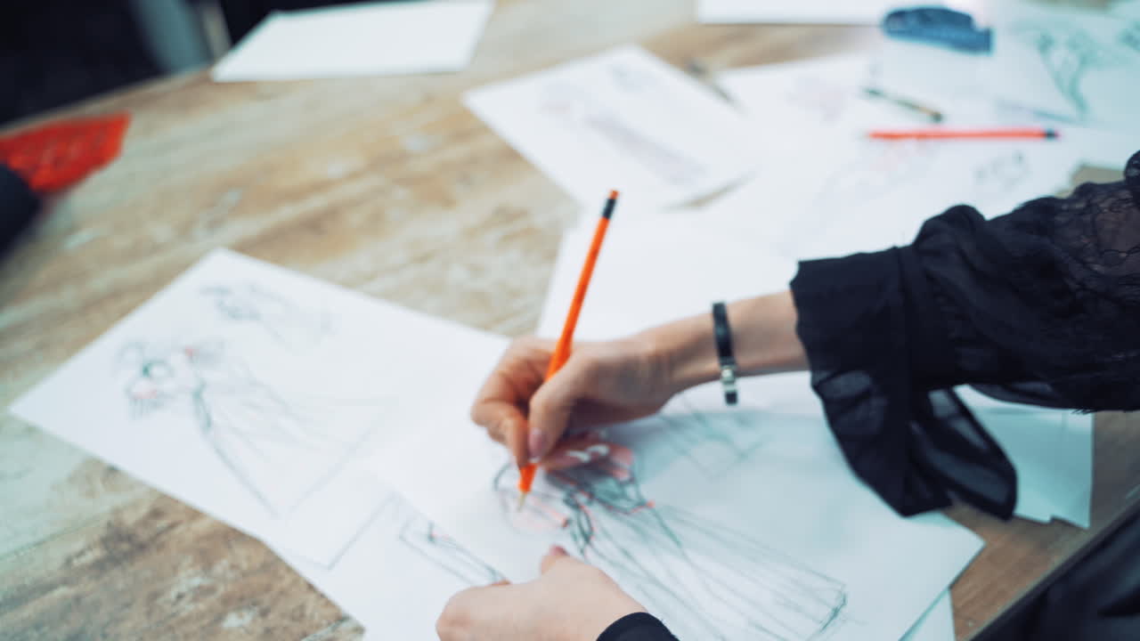 Process of drawing sketches of a model dress on papers by a female tailor. Professional designer draws illustrations for future dresses with a pencil on the table in atelier.