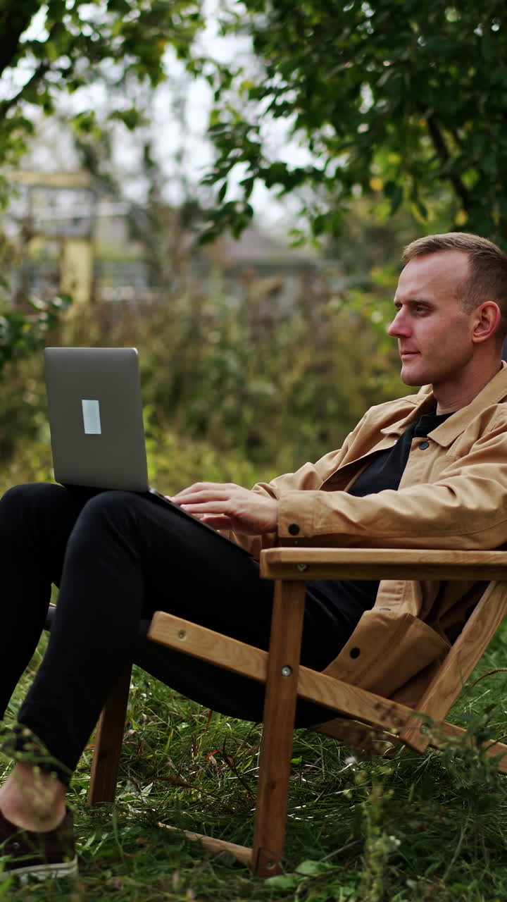 Busy man focused on his work on laptop sitting in garden. Camera slowly approaching freelancer who finishes work, closes laptop and leans back in his chair. Vertical video