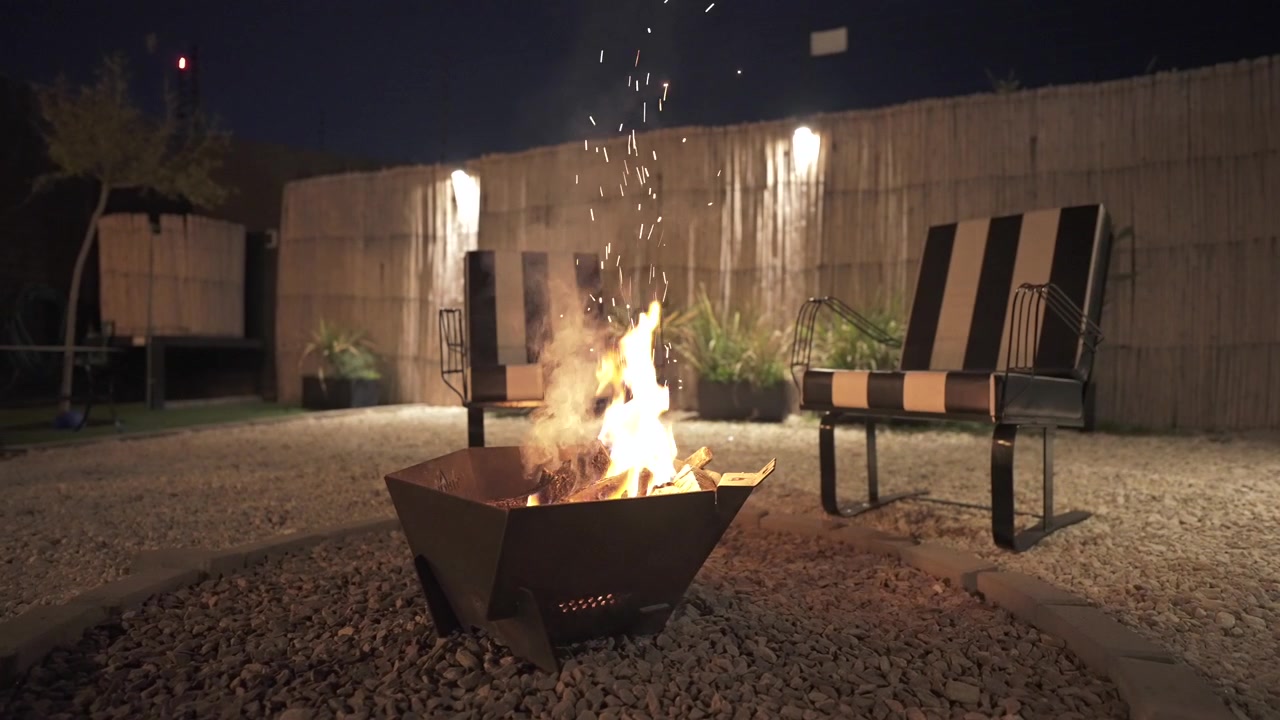Braciere in metallo con fiamme, affiancato da due moderne sedie a righe bianche e nere. Clip generale in slow motion che si muove leggermente a destra.