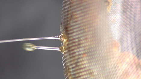 Sideways-perspective-view-of-clear-liquid-oil-dripping-down-from-metal-strainer-in-process-of-cooking-homemade-meal,-extreme-close-up-static-shallow-depth-of-field