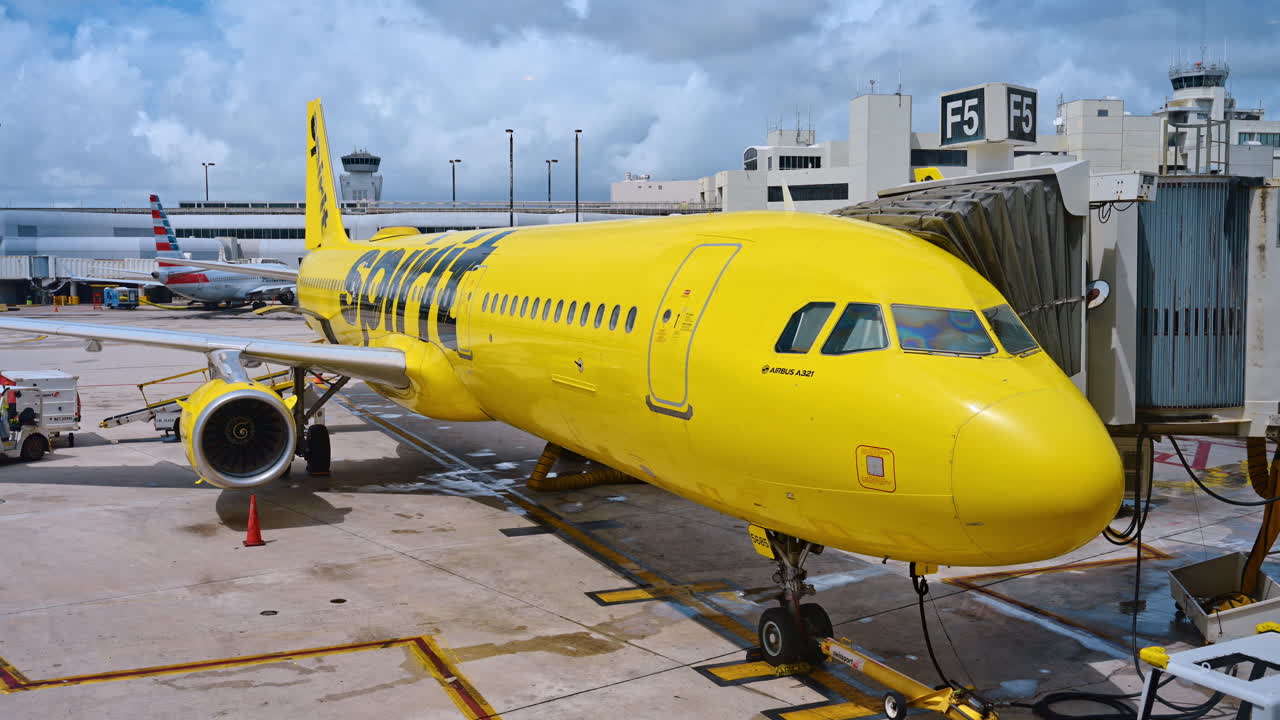 Miami, AS, 9 April 2026: Pesawat komersial kuning di gerbang bandara untuk boarding. Tampilan samping sebuah Airbus A321 kuning yang terhubung ke garbarata di terminal di bawah langit berawan.