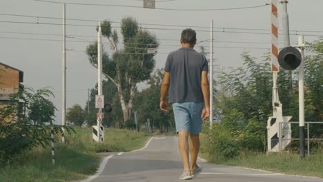 Shot-of-a-man-walking-in-the-countryside-seen-from-behind,-he-is-passing-over-the-tracks-of-an-open-railroad-crossing,-it-is-sunny-and-it-is-summer