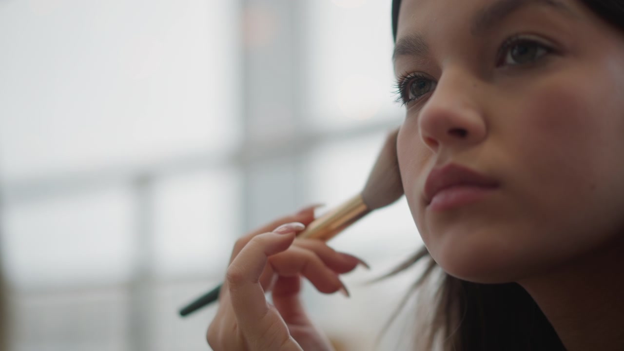 Close profile of young woman receiving powder makeup gently blended across cheek, jawline, and nose with fluffy brush; soft window light and calm expression support polished beauty routine