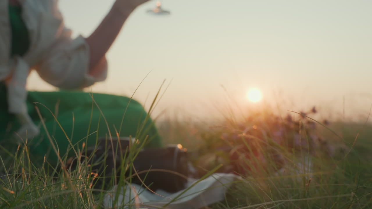 Woman seated on grassy hill reaches for wine glass, gazes into distant sunset over river then sips red wine, scenic picnic setup with fruit, flowers and camera in golden hour light