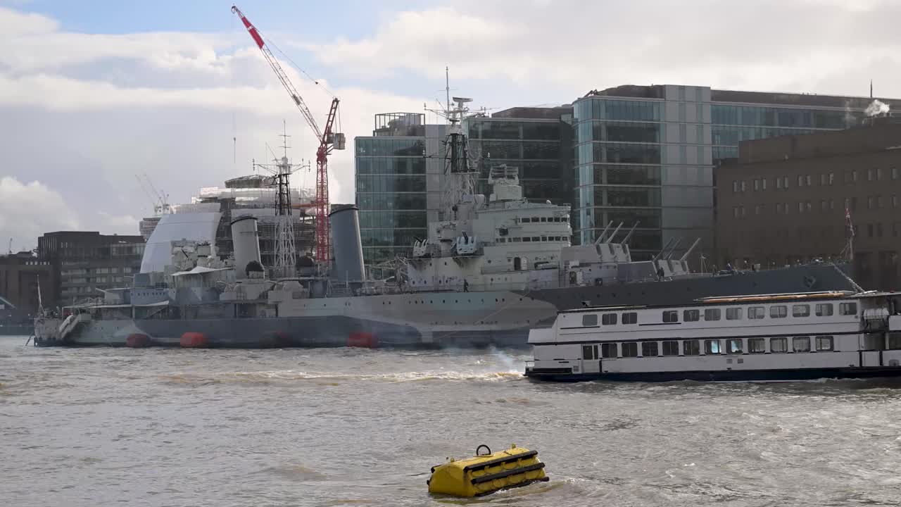 Varen over de rivier de Theems langs de enorme grijze romp en geschuttorens van het historische lichte kruisermuseumschip HMS Belfast, gelegen tussen London Bridge en Tower Bridge.