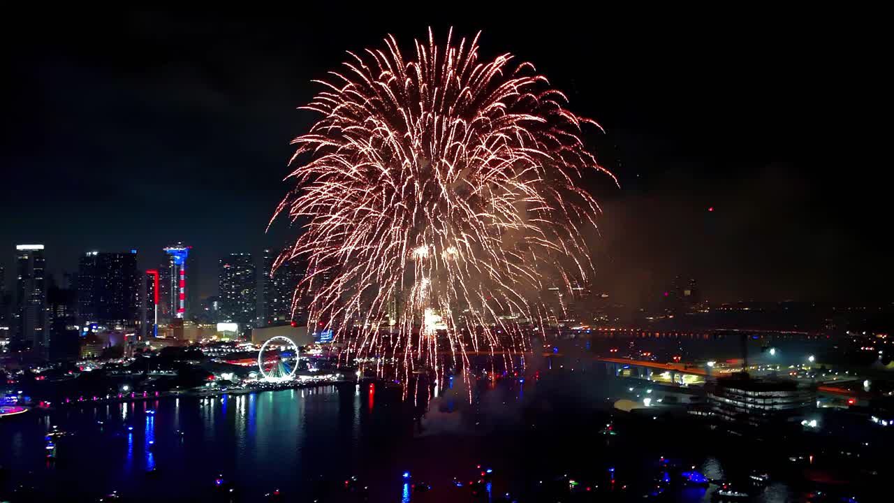 Elevated drone shot of fireworks bursting in brilliant patterns, mirrored in the bay with city lights sparkling in the distance