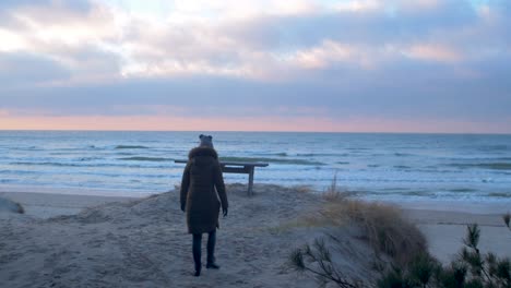 Mujer-Joven-Con-Ropa-De-Invierno-Camina-A-Lo-Largo-De-La-Orilla-Arenosa-De-La-Playa-Del-Mar-Báltico-Al-Atardecer-Romántico,-Banco-De-Madera,-Foque-Medio-Disparado