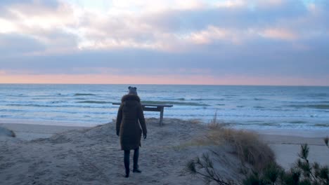 young woman in winter clothes walks along the sandy shore of the baltic sea beach at romantic sunset, wooden bench, medium jib shot up