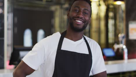 Retrato-De-Un-Barista-Afroamericano-Sonriendo-A-La-Cámara-Usando-Delantal-En-La-Cafetería