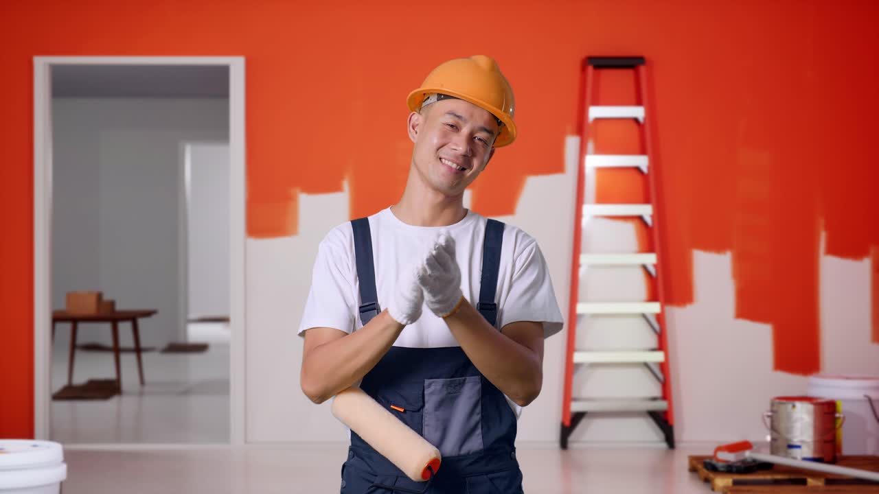 pintor hombre asiático con casco de seguridad sonriendo y aplaudiendo sus manos mientras está de pie con la pared de pintura en la habitación