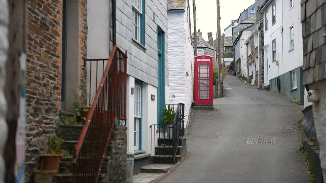 A street in Port Isaac, Cornwall, featuring a red British phone box positioned along a narrow uphill lane with tightly packed stone and whitewashed buildings in the village of Portwenn