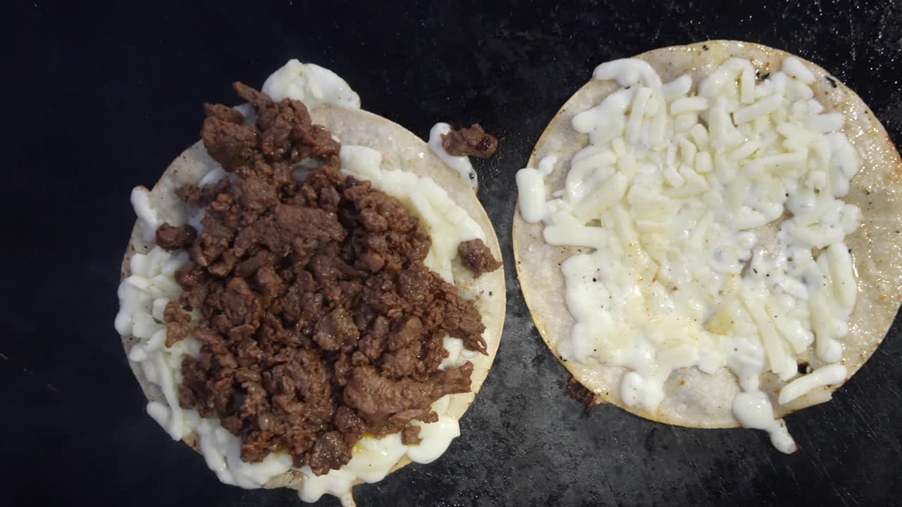 Næroptagelse af to tortillas, der damper på en varm grill, den ene fyldt med grillet asada og den anden med smeltet ost. Stigende damp understreger varmen, tilberedningsprocessen og forberedelsen af gadekøkken.