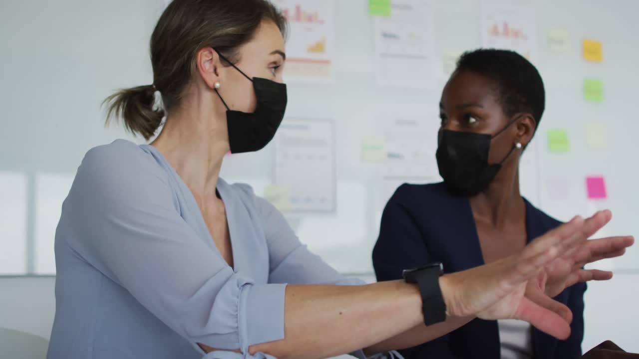 dos colegas femeninas diversas con máscaras faciales, sentadas en el escritorio y hablando