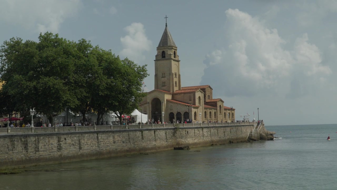 Udsigt til den ikoniske San Pedro Kirke langs strandpromenaden nær San Lorenzo-stranden i Gijon, Asturien, Spanien, på en lys dag med turister og dramatiske skyformationer på himlen.
