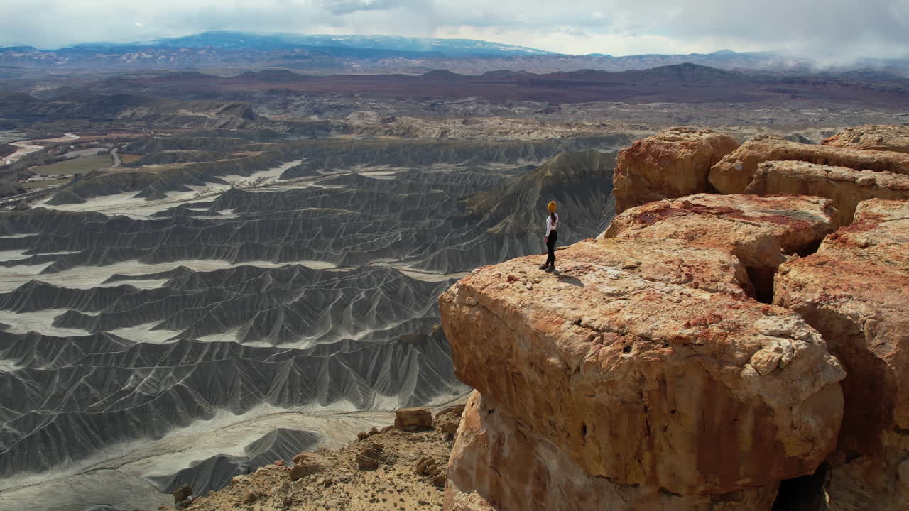 vista aérea de una mujer solitaria de pie en el borde de un acantilado y mirando el impresionante paisaje desértico, tiro de drone en órbita