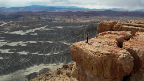 Vista-Aérea-De-Una-Mujer-Solitaria-Parada-En-El-Borde-Del-Acantilado-Y-Mirando-El-Impresionante-Paisaje-Desértico,-Orbitando-Un-Disparo-De-Drone