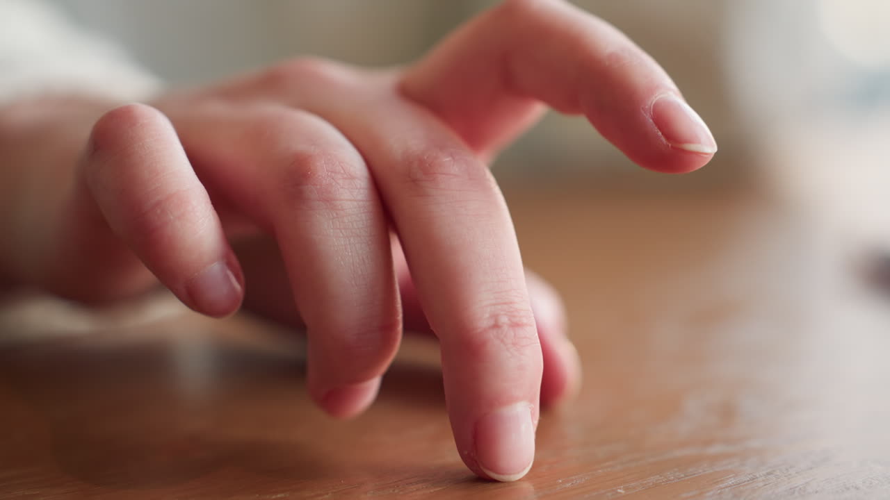 Close up hand of white person in white sleeve slowly tapping wooden table with finger during thoughtful moment in soft natural indoor light expressing calm, reflection