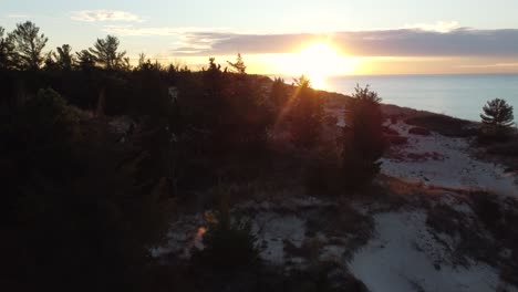 Aerial-dolly-over-snowy-forest-landscape-towards-setting-sun-by-water-shoreline