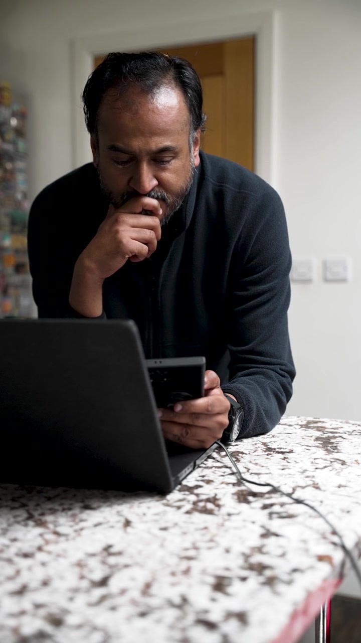 An adult man leans over a granite countertop using a laptop and smartphone while working remotely from home. vertical video