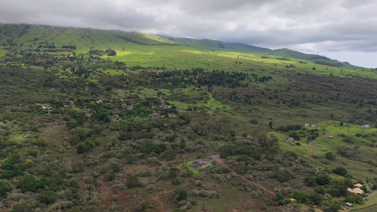 Ripresa aerea con drone 4K a 60fps dell'Upcountry Maui. Verdi colline lussureggianti e una vasta proprietà immobiliare rurale si estendono sotto nuvole basse, con la luce del sole che illumina il vibrante paesaggio montano tropicale