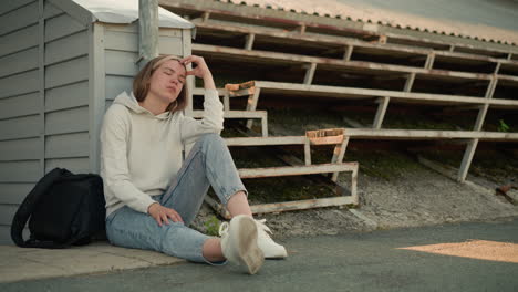 lady in jeans seated with hand resting on head, deep in thought, in stadium setting, black bag beside her, surrounded by rustic bleachers