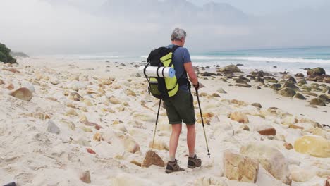 senior hiker man with backpack and hiking poles walking while hiking on the beach.