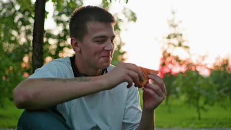 man with tattooed hand making a toast with bread, with a partial view of another person holding bread in the background, sunlight filters through trees, creating a warm outdoor atmosphere