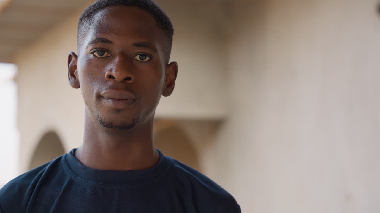 Ritratto di uomo di colore in corridoio, giovane uomo africano sotto un passaggio ad arco, leggero sudore sulla fronte, sguardo contemplativo ed espressione naturale, luce solare soffusa che crea un caldo contrasto tonale, profondità di campo ridotta