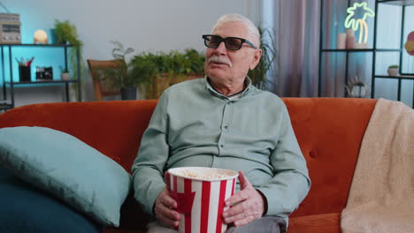 Abuelo-En-El-Sofá-Comiendo-Palomitas-De-Maíz-Y-Viendo-Interesantes-Series-De-Televisión,-Juegos-Deportivos-En-Línea-En-Casa