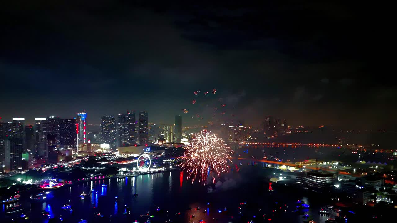 Stunning fireworks explode in radiant color while the drone hovers above, revealing glowing reflections shimmering across the night waters