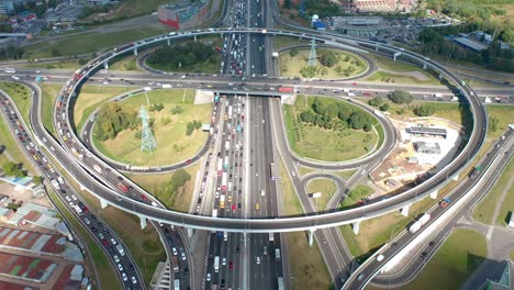 the large circular intersection with rounds and curve roads and clover exits. top down aerial view from drone. cars in typical traffic routine. sunlight with shadows of clouds. green areas and buildings all around. camera is moving forward over highway.