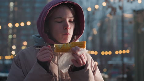 young woman enjoying maize held by stick wrapped in white sachet, steam rising from fresh corn with bokeh lights in blurred background, people walking in outdoor park setting during winter