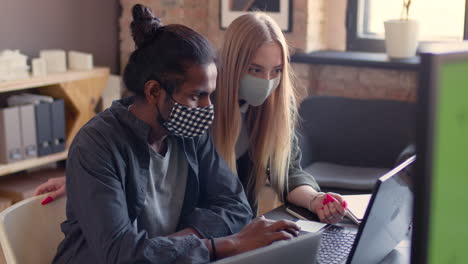 two multiethnic colleagues with face masks looking at laptop screen and discussing a new project in an animation studio