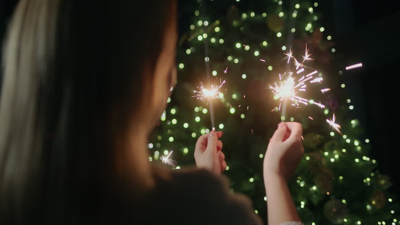 mujer feliz sosteniendo chispas, de pie contra el telón de fondo de un árbol de navidad en casa. vista trasera