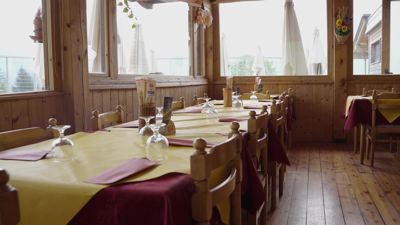 Wide interior shot of a welcoming alpine lodge dining hall in Trentino, Italy. Long wooden tables ready for guests, with large windows framing mountain views