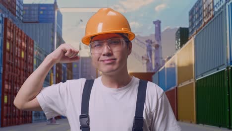 close up de un hombre asiático que trabaja con gafas de protección y casco de seguridad sonriendo y flexionando su bíceps mientras está de pie en el almacén del patio de contenedores