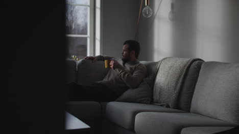 Man-sitting-on-couch-and-drink-coffee-in-moody-winter-season,-side-view