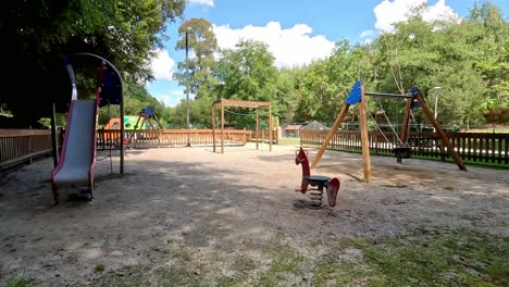 general-view-of-the-playground-empty-without-children-with-swing-moving-alone-and-slide,-sunny-day-with-clouds-in-the-middle-of-the-trees,-shot-blocked,-Ordes,-Galicia,-Spain